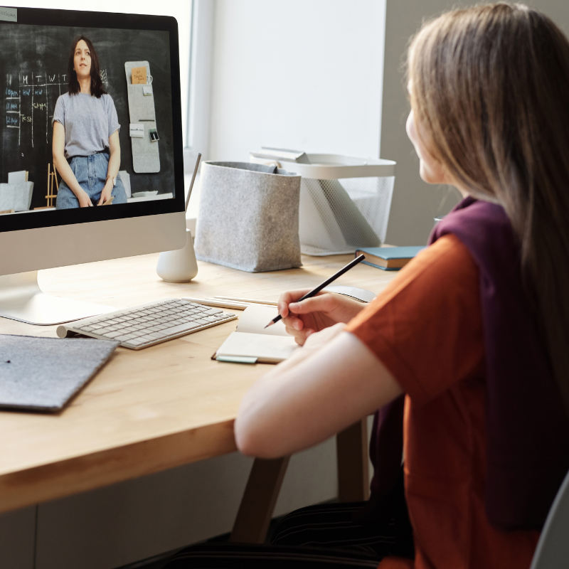 Embedded website video viewed on a desktop monitor, showing a woman teaching online while a viewer takes notes at their desk.