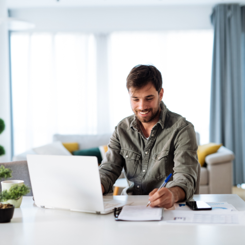 Man creating online courses, smiling while taking notes and working on a laptop at home.