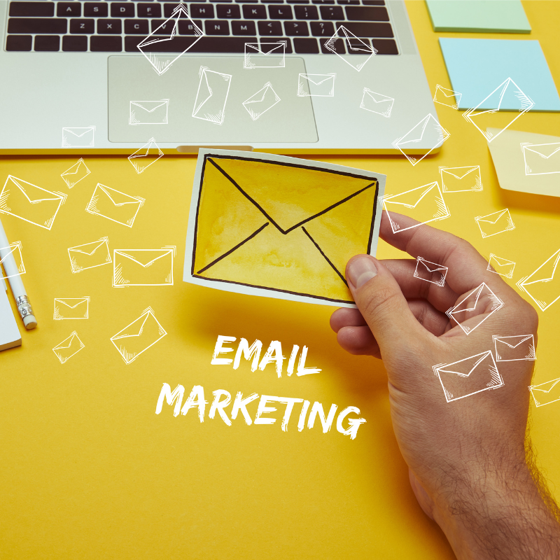 Hand holding an envelope icon in front of a laptop with “Email Marketing” text on a yellow background, representing a digital email campaign.