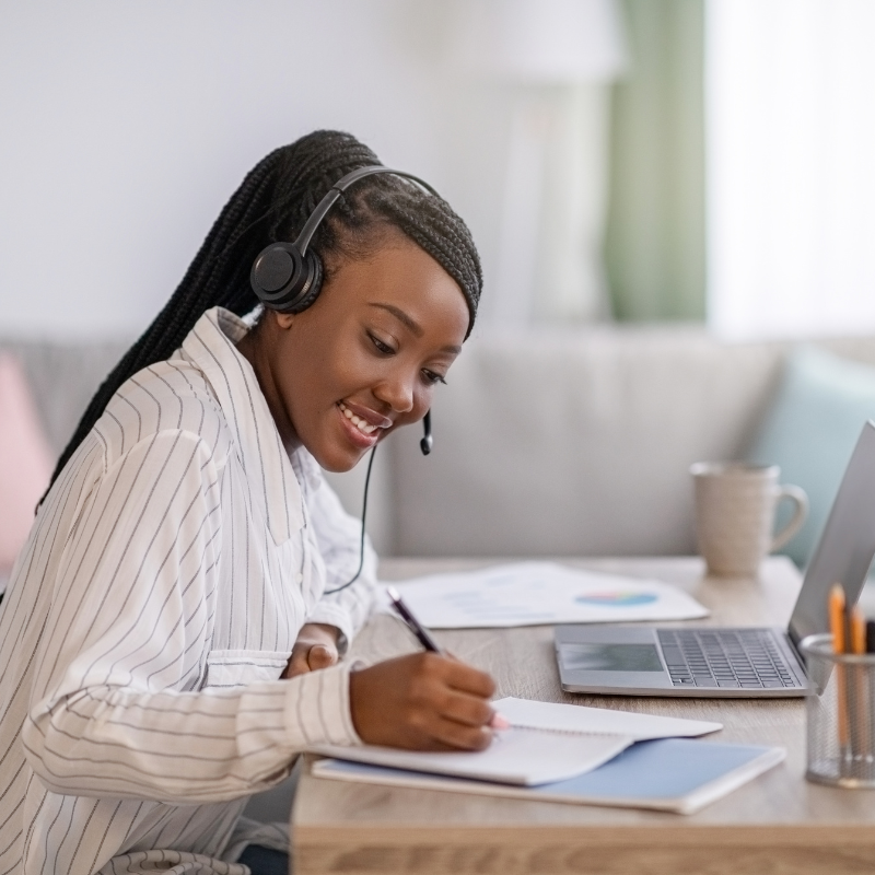 More than an online course concept shown by a smiling woman taking notes during a virtual learning session with a headset and open laptop.